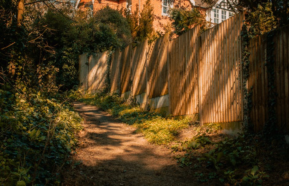 A narrow dirt garden path with uneven, compacted soil, bordered by lush green shrubs and low-growing plants on the left side, leading alongside a tall wooden fence composed of vertical slats. The fenc