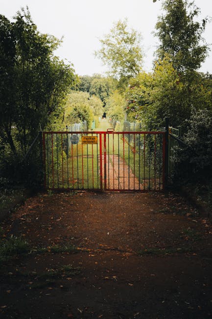 A narrow dirt garden path with uneven, compacted soil, bordered by lush green shrubs and low-growing plants on the left side, leading alongside a tall wooden fence composed of vertical slats. The fenc