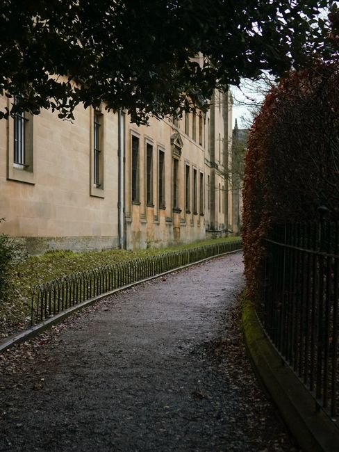A narrow, winding gravel pathway in a garden setting runs alongside a historic stone building with multiple tall, rectangular windows, surrounded by lush, trimmed hedges and mature trees. The pathway 